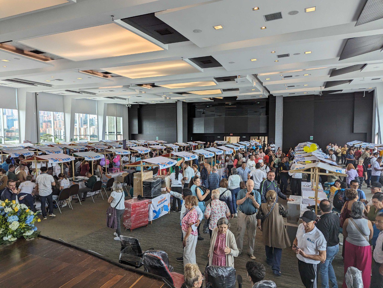 Import resale cross-border EC is a large-scale indoor event where a large number of people stand and converse around a table with various items on display in a room with large windows and views of the city.